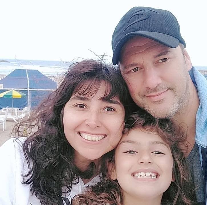 Familia de tres sonriendo para una selfie en la playa con una carpa azul detrás.
