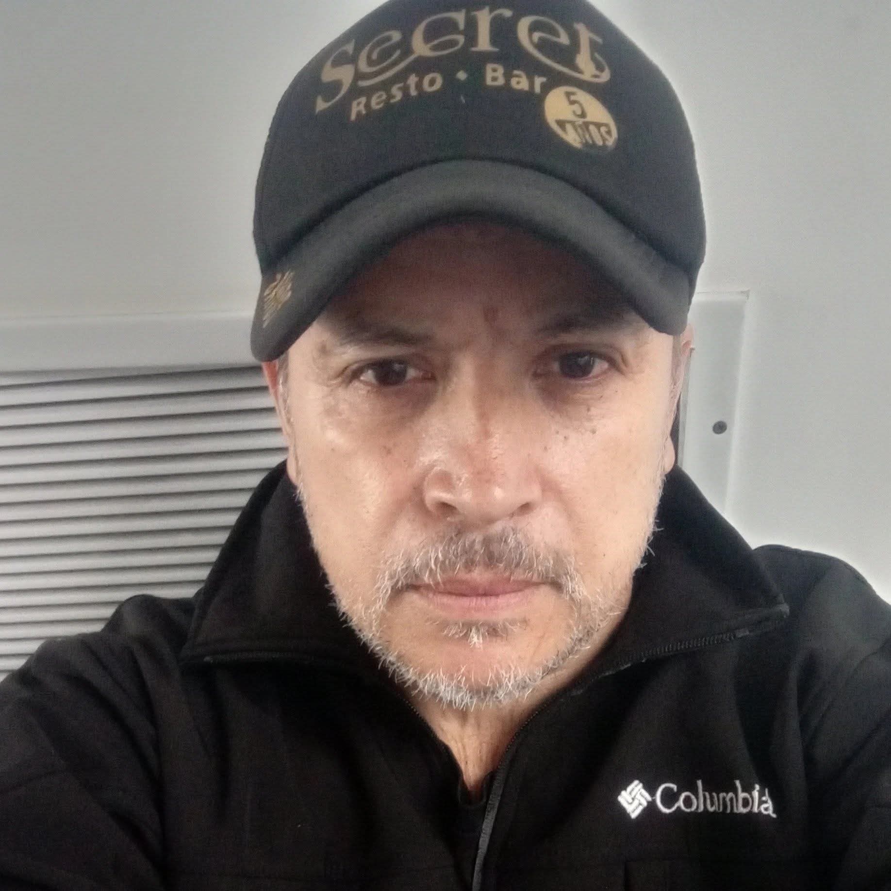 Hombre con barba canosa, gorra negra de Secret Resto Bar y chaqueta negra marca Columbia.