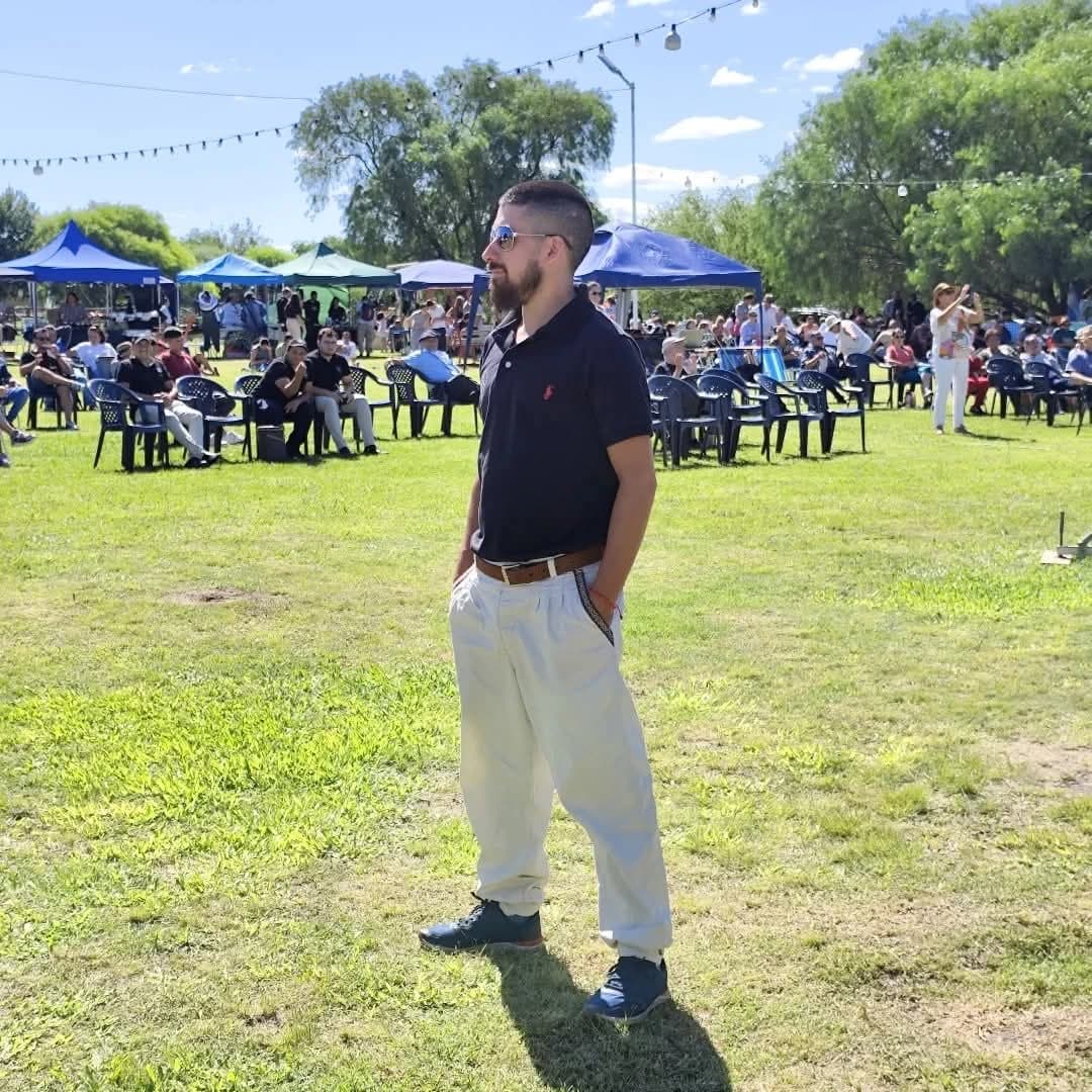 Hombre con polo negro y pantalones claros parado en un campo con carpas al fondo.