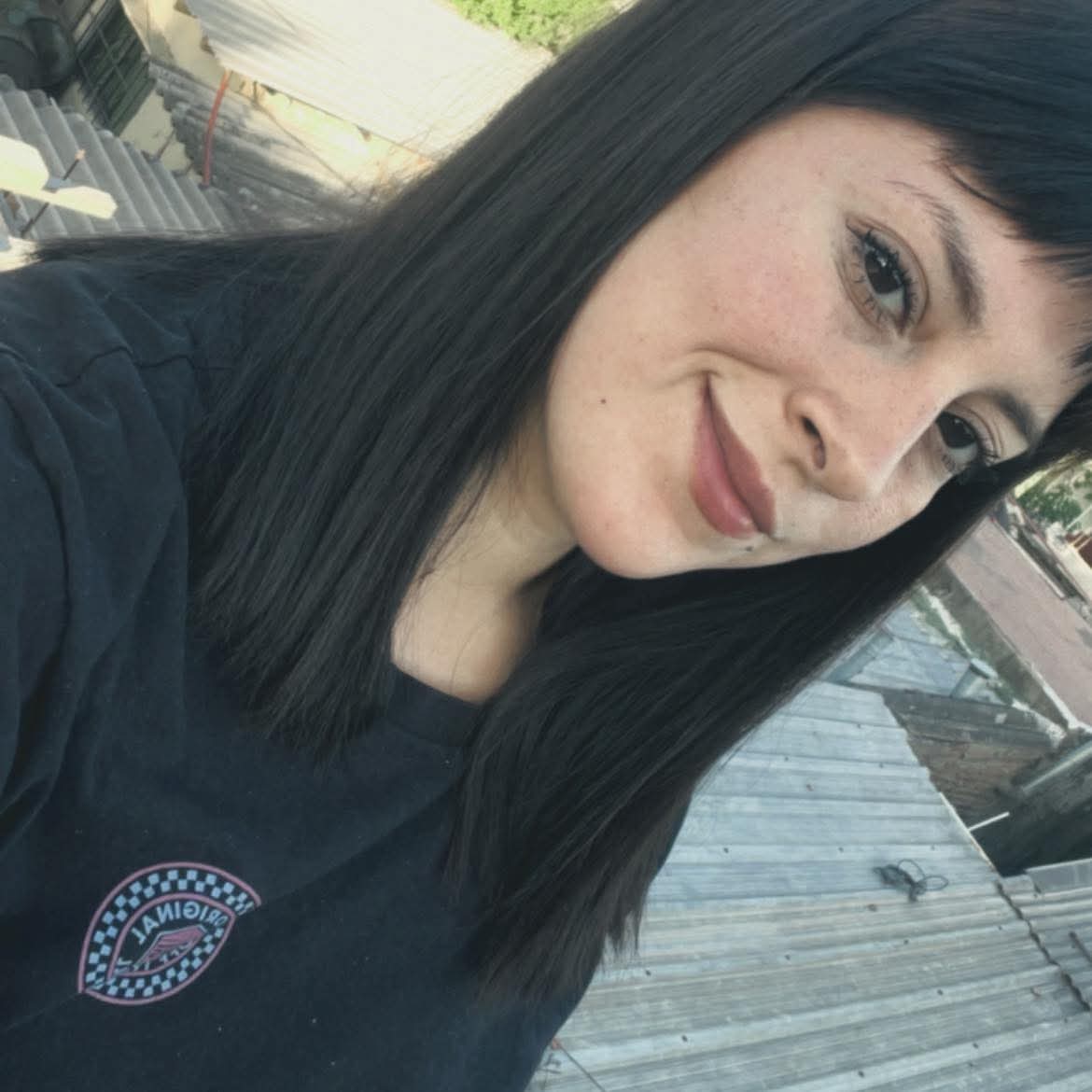 Mujer joven con pecas y cabello negro liso sonriendo frente a un techo metálico.